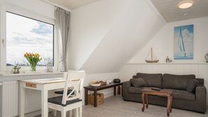A cozy living room with a white desk, a brown sofa, and a wooden coffee table. A window with a view of the sky and a sailboat painting on the wall. - Ferienwohnungen Haus Schau ins Land (Emmelsbuell-Horsbuell)