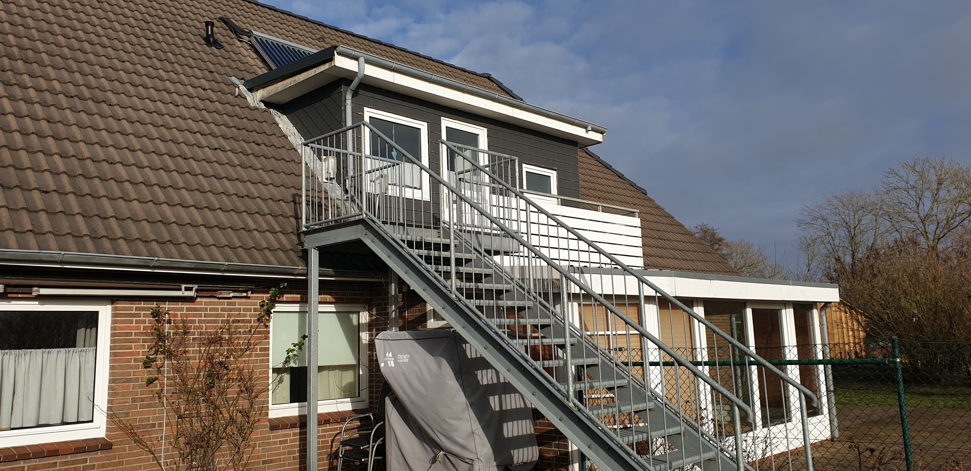 A residential house with a brick facade, a metal staircase leading to a balcony, and a solar panel on the roof.