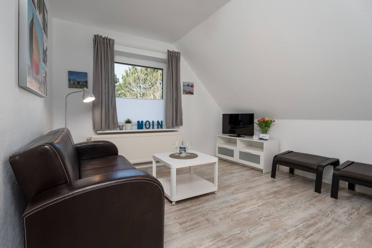 Un salon moderne avec un canapé en cuir marron, une table basse blanche, un téléviseur posé sur un meuble blanc et une fenêtre avec des rideaux.