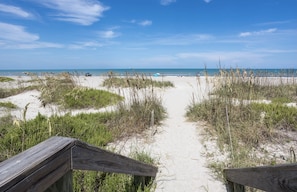 Un sentier de sable qui longe la plage et mène à l’océan, bordé d’herbe et d’une clôture en bois.