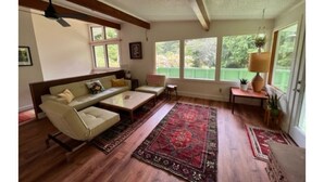 A spacious living room with a sofa, coffee table, and a large window providing a view of greenery outside. - Private mid-century modern hideaway in forest near the river and sea. (Crescent City)