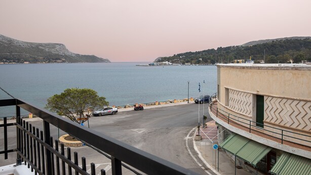 A coastal view from a balcony overlooking a road leading to the sea, with buildings and a tree in the foreground. - White Heaven. Near the Marina Lakki and a Wonderful View of the Port. (Leros)