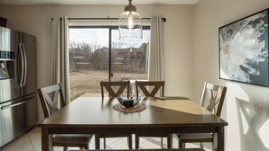 A dining area with a wooden table, four chairs, a hanging light fixture, a refrigerator, and a window with curtains. - Jenks Spectacular Spain Ranch-Close to Five Oaks (Jenks)