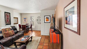 Un salón con un sofá de cuero, una mesa de centro y un mueble para televisión con una manta. Hay cuadros en las paredes y una zona de comedor con sillas.