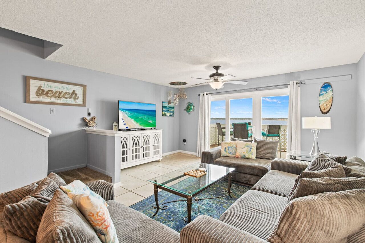 A living room with a sofa set, a coffee table, a TV, and a view of the beach through large windows.