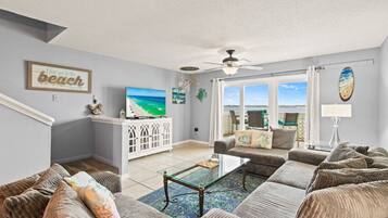 A living room with a sofa set, a coffee table, a TV, and a view of the beach through large windows.