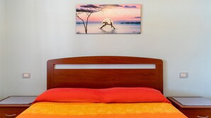 A bedroom with a wooden bed, a red and orange bedspread, and a painting of a sunset over water on the wall. - Su Castangiu1, flat in villa with swimming pool and 5-a-side football pitch (tertenia)