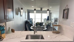 A modern kitchen with a marble countertop, stainless steel sink, and dark cabinetry. There is a dining area with a round table and chairs in the background. - Galveston Oceanview,Wonderful location! 2bed/2bath (Galveston)