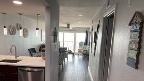 A modern kitchen with a stainless steel dishwasher, a dining area with chairs, and a hallway with a ceiling fan and decorative wall art. - Galveston Oceanview,Wonderful location! 2bed/2bath (Galveston)