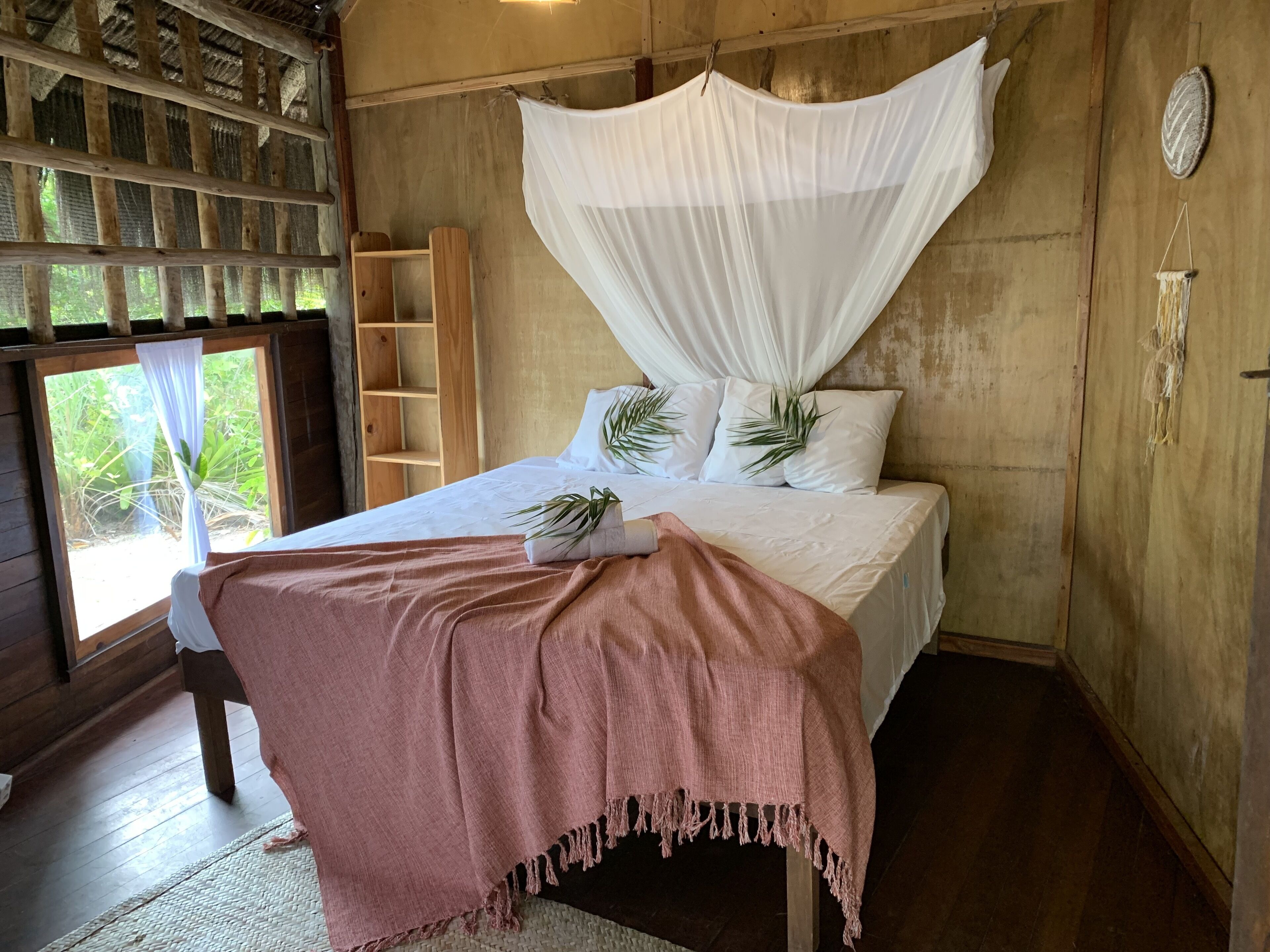 Quarto com cama, dossel, prateleira de madeira e janela com vista para o verde.