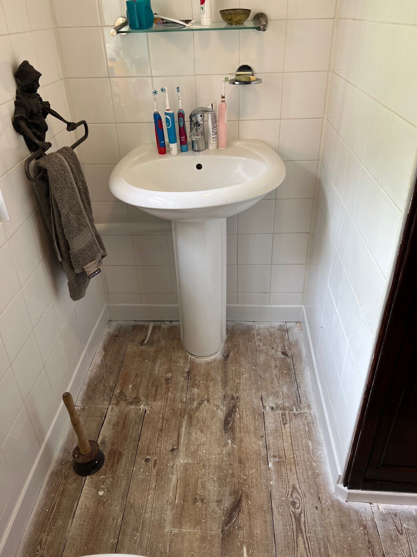 Casa de banho com lavatório pedestal branco, chão em madeira, suporte de toalhas com toalha e escovas de dentes numa prateleira.