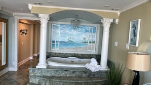 Une salle de bain dotée d’une grande baignoire, offrant une vue sur la plage grâce à une fenêtre ouverte, et agrémentée d’un lustre.