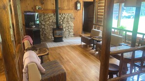 Un interior acogedor de cabaña con una pared de piedra, pisos de madera, una chimenea y un comedor con sillas y una mesa.