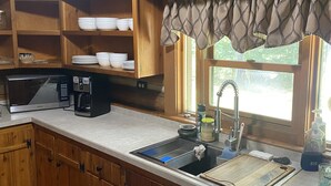 Una cocina con armarios de madera, microondas, cafetera y ventana con cortina.