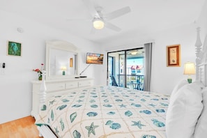 A bedroom with a bed, a ceiling fan, a TV, a balcony, and a view of the outside.