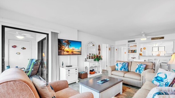 A spacious living room with a large flat-screen TV, a sofa set, a coffee table, and a dining area with a kitchen in the background. - Runaway Bay 283 by Duncan Real Estate (Bradenton Beach)