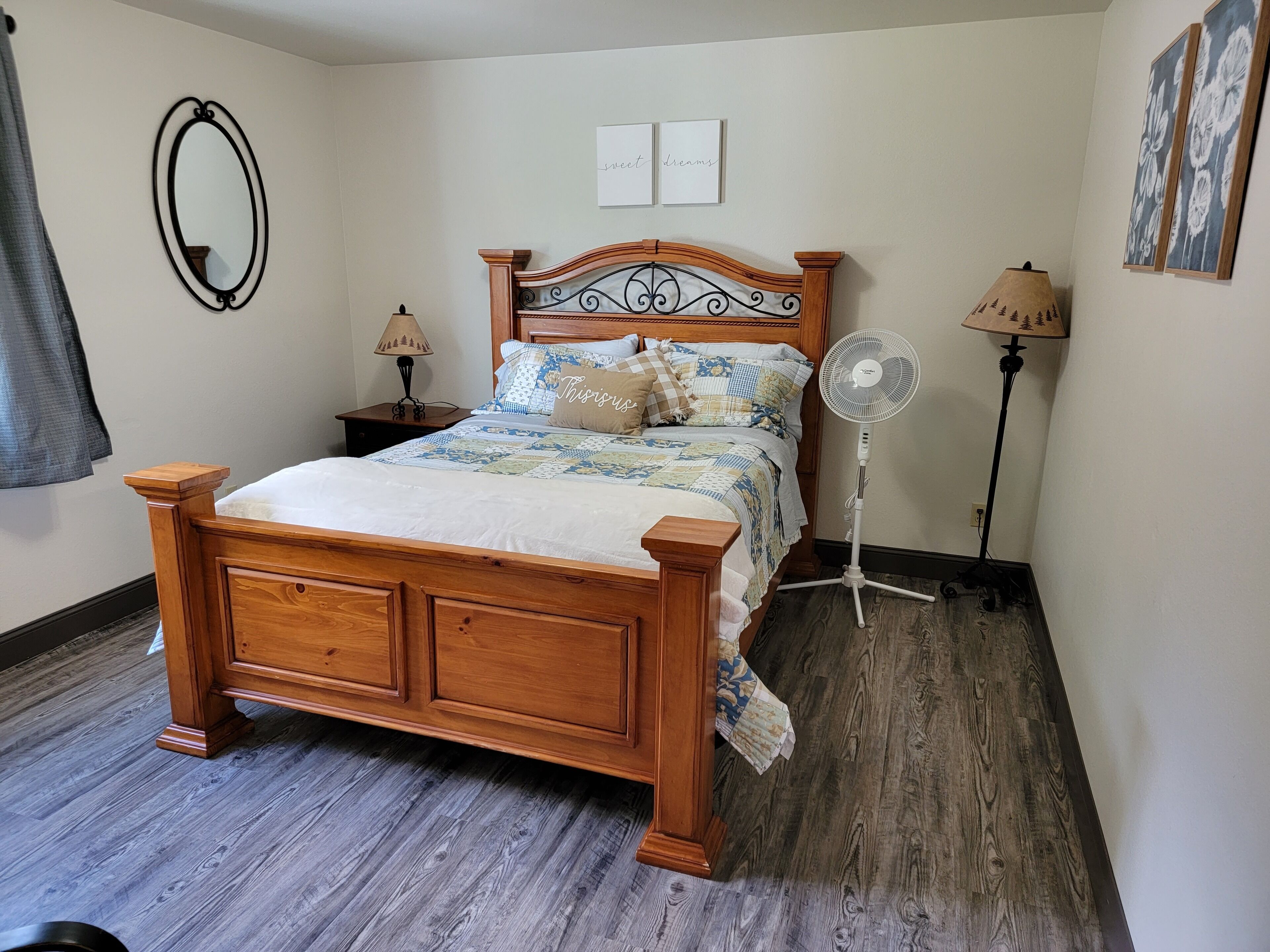 Un lit en bois avec une tête de lit, une table de chevet avec une lampe, un ventilateur sur pied et un miroir fixé au mur.