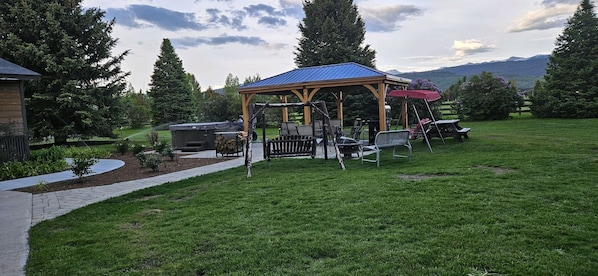 A gazebo with a blue roof, outdoor seating, and a barbecue grill in a grassy backyard with trees and mountains in the background.