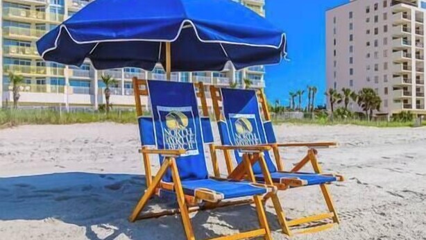 Dos sillas de playa con una sombrilla azul sobre la arena, con edificios altos al fondo.
