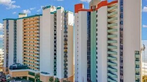 A row of high-rise residential buildings with balconies, a clear sky, and a parking area in the foreground.