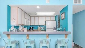 A kitchen with a blue accent wall, white cabinets, a countertop, and three bar stools. - Stay on the Boardwalk | Oceanfront w/ Pools! (Myrtle Beach)
