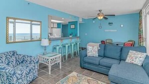 A living room with a blue sofa, a patterned armchair, a small white table, and a ceiling fan. - Stay on the Boardwalk | Oceanfront w/ Pools! (Myrtle Beach)
