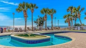A swimming pool with a curved design, surrounded by palm trees and lounge chairs, with a view of the ocean in the background. - Stay on the Boardwalk | Oceanfront w/ Pools! (Myrtle Beach)