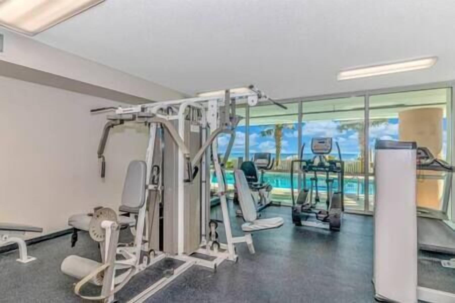 Un gimnasio con diversas máquinas de ejercicio, un ventanal con vista a una piscina e iluminación empotrada en el techo.
