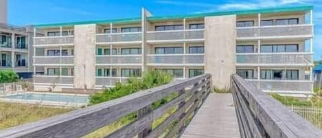 A multi-story hotel with balconies, a wooden boardwalk leading to the entrance, and a clear blue sky.