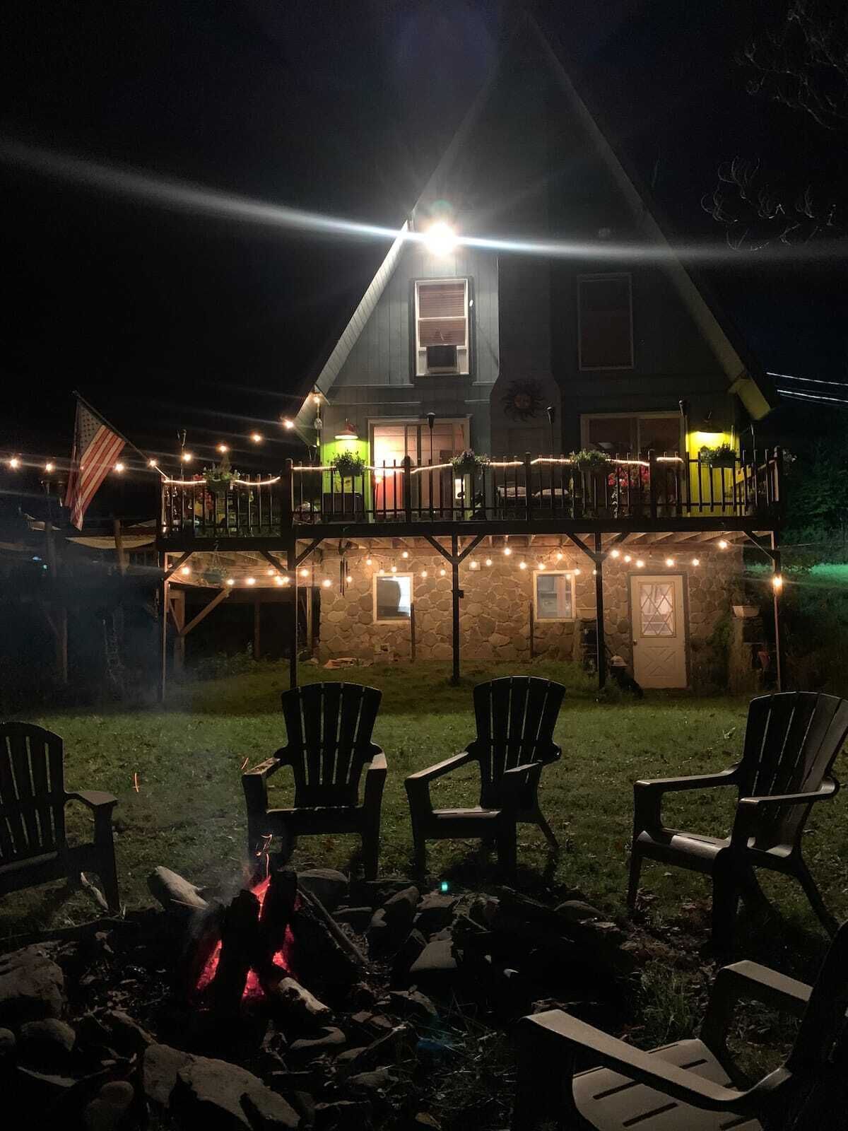 Una casa de dos pisos con balcón, luces colgantes y una zona con fogata y sillas.