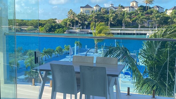 Un salón moderno con pared de vidrio que da a una piscina y una vista panorámica de la costa, con casas y palmeras.