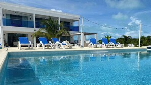 Una piscina cristalina con sillas reclinables azules y un edificio moderno con ventanas de tono azulino al fondo.