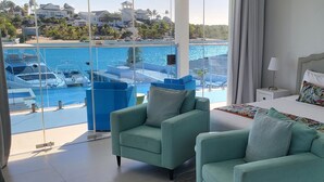 Habitación de hotel con un gran ventanal que da a un puerto deportivo y una piscina. Hay dos sillones color turquesa y una cama con un cabecero llamativo.