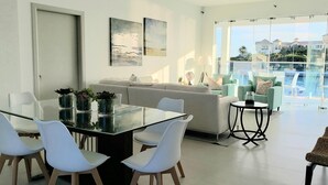 Una sala moderna con una mesa de vidrio, sillas blancas, un sofá y un balcón con vistas a casas y palmeras.