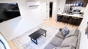 A modern living room with a flat-screen TV, a sofa with patterned cushions, a coffee table, and a kitchen area with a refrigerator, microwave, and bar stools.