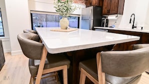 Een moderne keuken met een marmeren aanrecht, houten stoelen, een bakstenen muur en uitzicht op het exterieur.