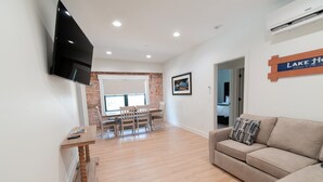 A modern living room with a flat-screen TV, a wooden dining table with chairs, a beige sofa, and a sign reading "LAKE HOUSE."