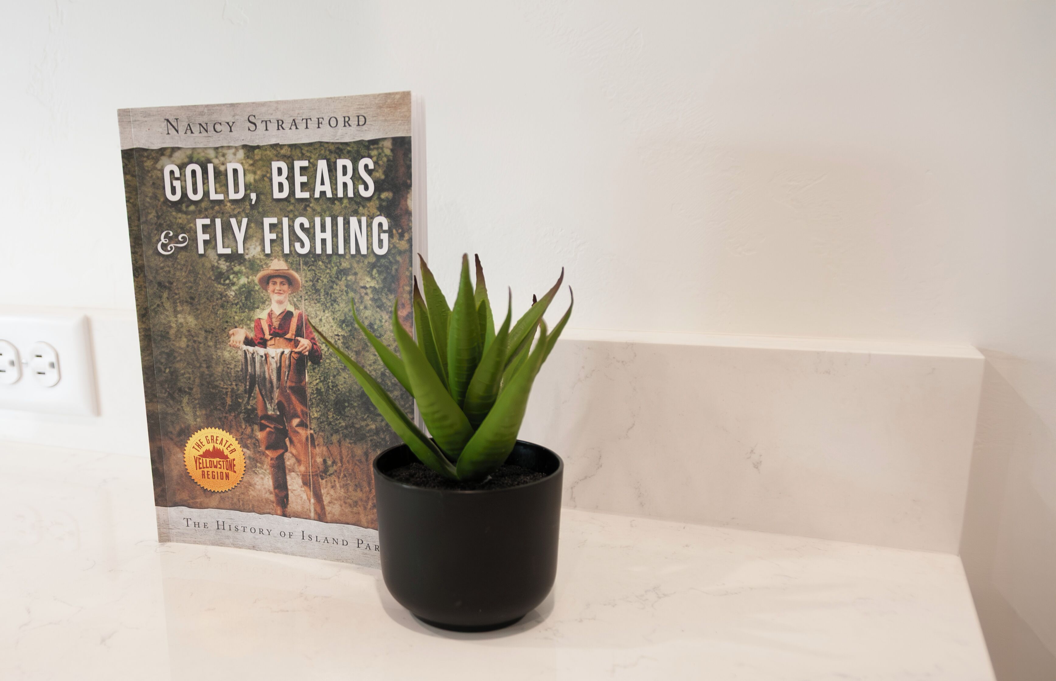 A book titled 'Gold, Bears & Fly Fishing' by Nancy Stratford placed next to a potted plant on a white surface.