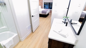 A modern hotel room with a wooden floor, a white bathtub, a large mirror, and a bed with a brick wall backdrop.