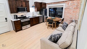A compact kitchen with dark wooden cabinets, a stainless steel refrigerator, and a dining area with a brick wall and large window.