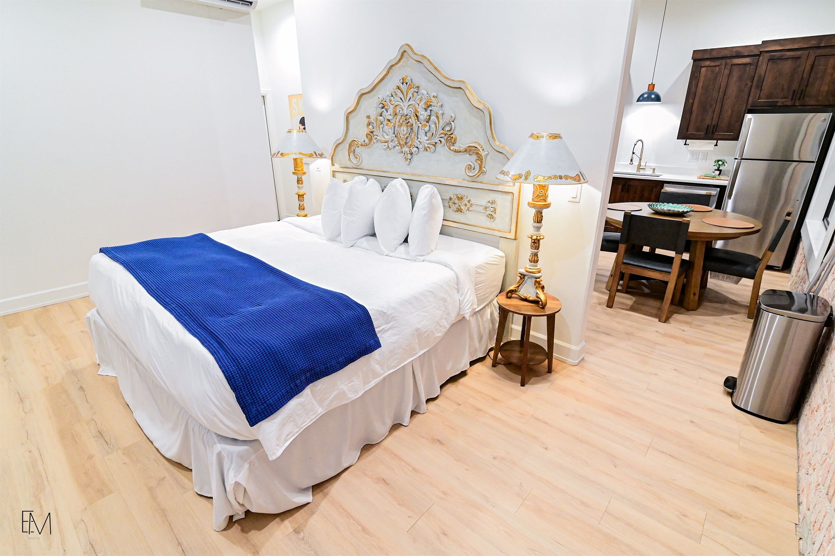 Une chambre avec un grand lit, une table de chevet avec une lampe, un petit tabouret, une poubelle et, au fond, un coin cuisine.