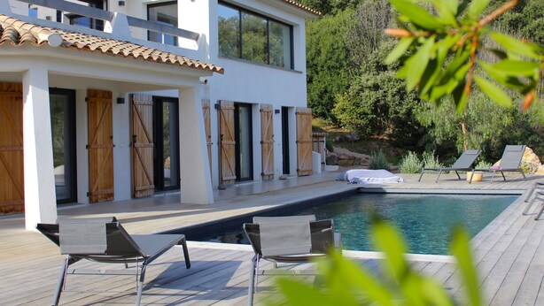 Une maison moderne à deux étages, dotée d’une piscine, de volets en bois et d’un espace salon extérieur.