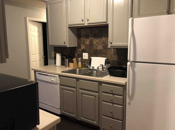 Kitchen area with a dishwasher, refrigerator, and sink, featuring a tiled backsplash and a door to an adjoining room.