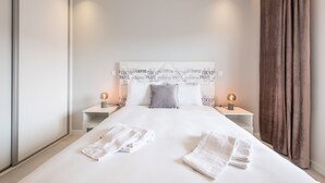Une chambre d’hôtel avec un grand lit, deux tables de chevet avec des lampes, une tête de lit ornée de noms de villes et un rideau sur le côté droit.