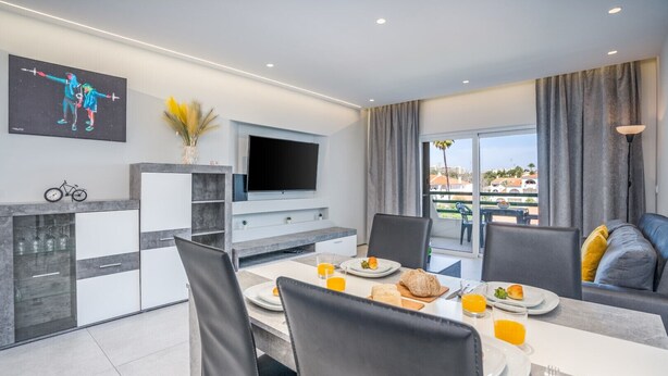 Un espace repas moderne doté d’un téléviseur à écran plat, d’une table à manger dressée avec des assiettes, des verres et un panier de fruits.
