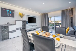 Un espace repas moderne doté d’un téléviseur à écran plat, d’une table à manger dressée avec des assiettes, des verres et un panier de fruits.