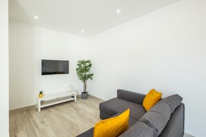 Un salon moderne avec un canapé gris, un banc blanc, un téléviseur à écran plat, une plante en pot et un petit ananas posé sur le banc.