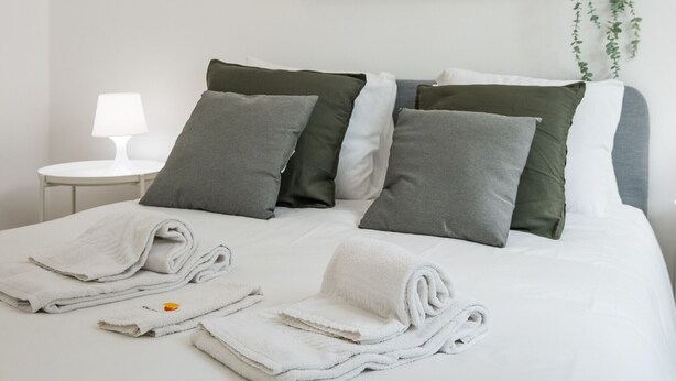 Una cama bien hecha con sábanas blancas y almohadas grises, una mesita de noche con lámpara y un estante en la pared con adornos.