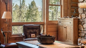 L’intérieur d’un chalet rustique, avec un fauteuil en cuir, une cheminée en pierre et une grande fenêtre donnant sur une forêt.