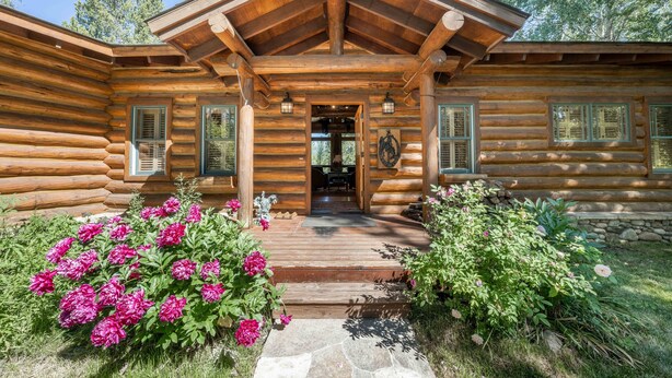 Une cabane en rondins avec une véranda en bois, des marches en pierre et un jardin parsemé de fleurs roses.